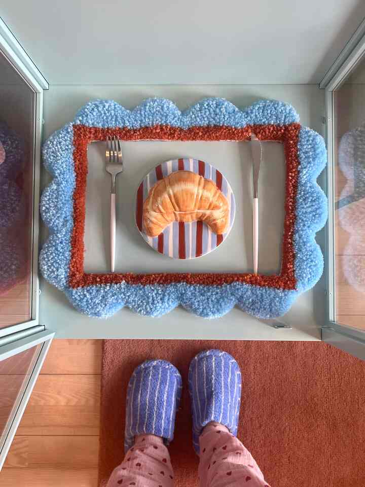 Simple kitchen scene featuring a light blue and brown rug, a croissant on a striped plate, and pink-handled cutlery