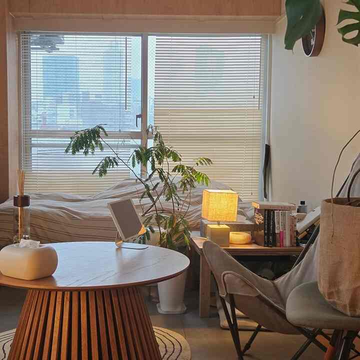 ナチュラルカラーと木目調が調和した一人暮らしの寝室、丸テーブルやストライプの掛け布団カバー、ブラインドが配置された雰囲気のあるインテリア