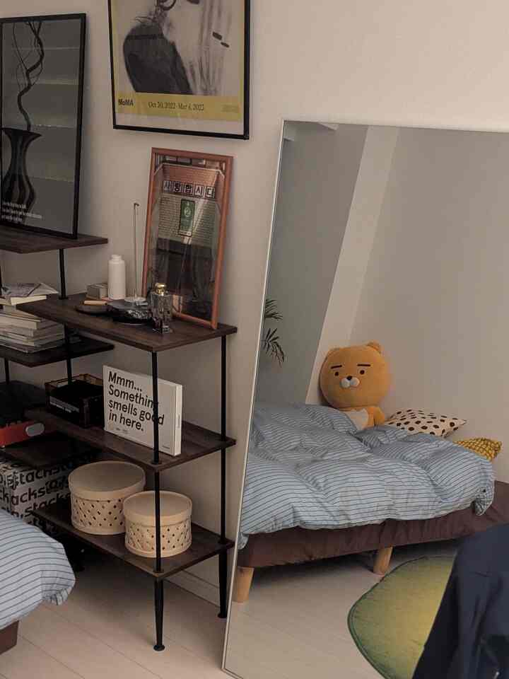 ナチュラルな色調の寝室、大きな鏡と木製の本棚が配置され、スカイブルーのボーダー掛け布団カバーが印象的な雰囲気のあるインテリア