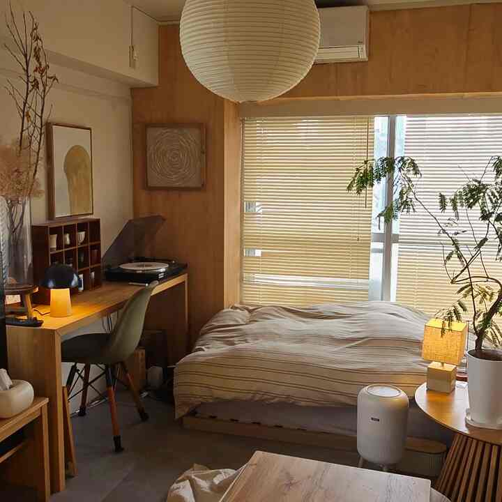 ナチュラルな木目調とベージュ色調の8畳寝室、デスクや植物、ブラインドが配置された落ち着いた一人暮らしの空間