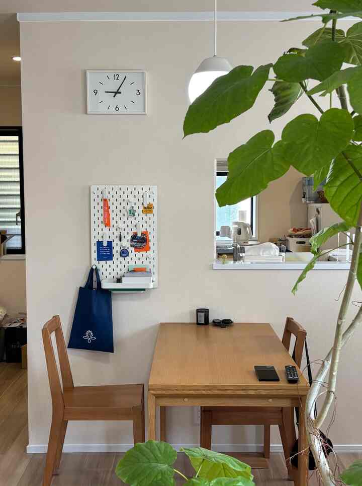 白い壁と茶色の木製家具が配置された二人用ダイニング、壁時計とペグボードメモパネルがある自然な雰囲気の空間