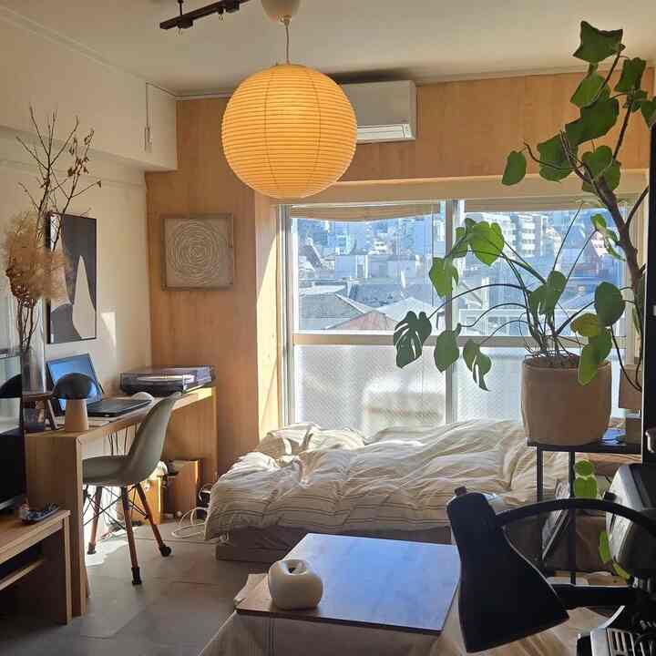 暖かみのある木目調と白い壁のワンルーム寝室、デスクと大きなペンダントライト、そして大きな観葉植物が配置された明るく居心地の良い空間