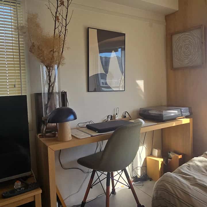 Natural-toned studio bedroom featuring a wooden desk, office chair, abstract poster, and table lamp in a minimal and cozy home office setup