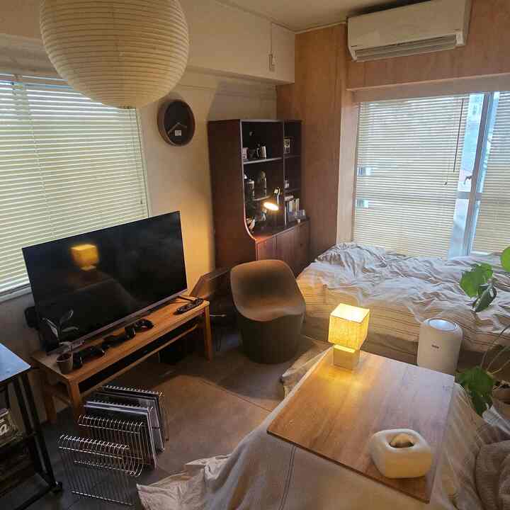 ナチュラルな色調のコンパクトなワンルーム寝室、中央に大きなペンダントライト、テレビ台、キャビネット、横に配置されたベッドとスツールがある印象的な空間