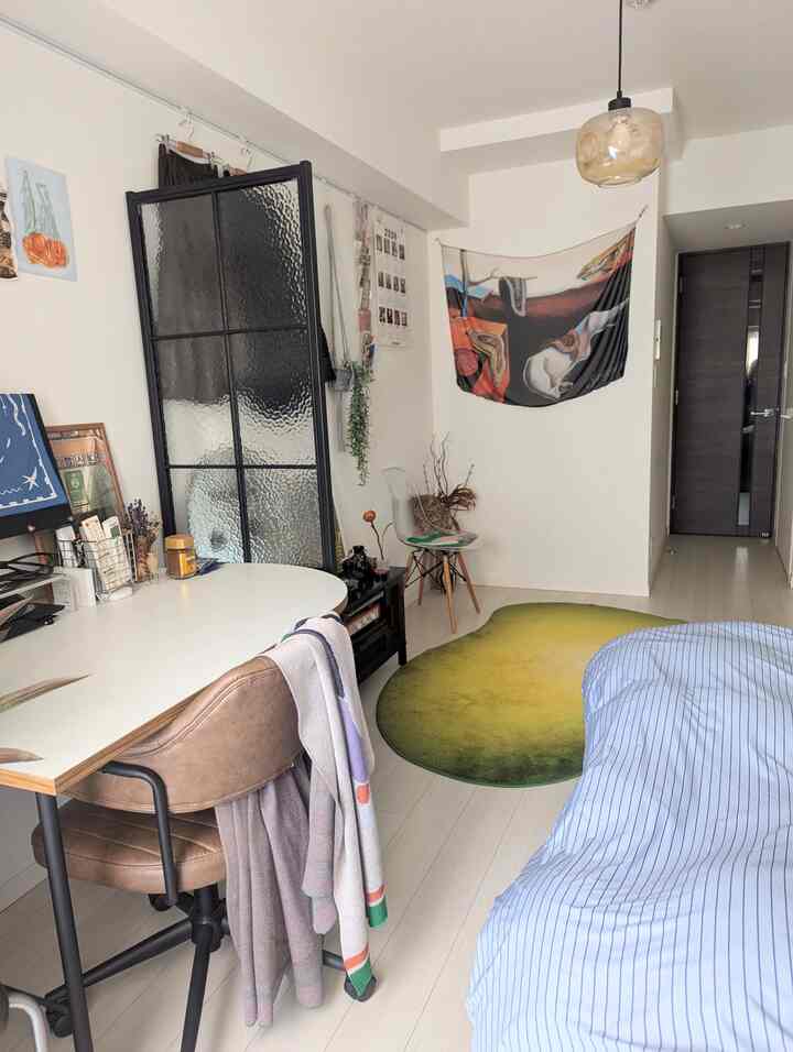 Studio apartment with white walls and floors, featuring a green gradient rug, a transparent chair, and a minimal desk setup