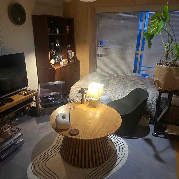 Studio apartment living room with warm wood tones and green accents featuring bed, round table, and blinds in a cozy natural modern style