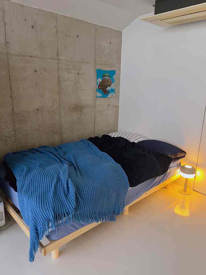 ナチュラルウッドトーンとコンクリート壁が調和するシンプルでミニマルな寝室、青いスローと小さなテーブルランプが配置された印象的な空間