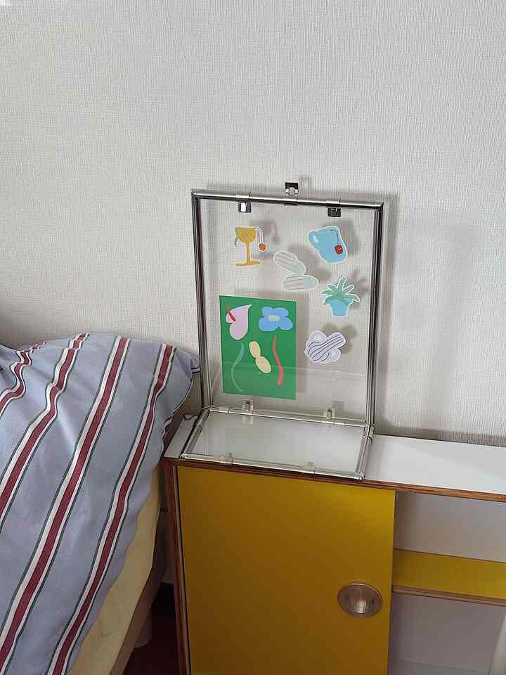 白い壁に沿って黄色いモジュール棚の上に透明な額縁が置かれたシンプルな寝室の一角