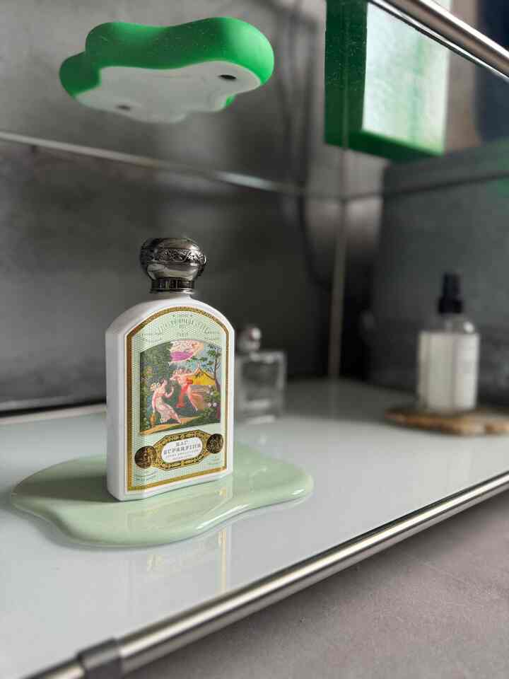 白と緑のトーンが特徴のバスルームの棚に、コースターの上に置かれたヴィンテージスタイルのボトルとディフューザーが配された落ち着いた印象のインテリア