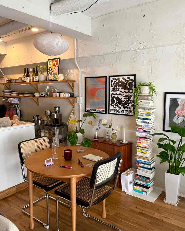A cozy dining room with white walls and wood-toned furniture featuring a round table and rattan chairs, decorated with posters and plants