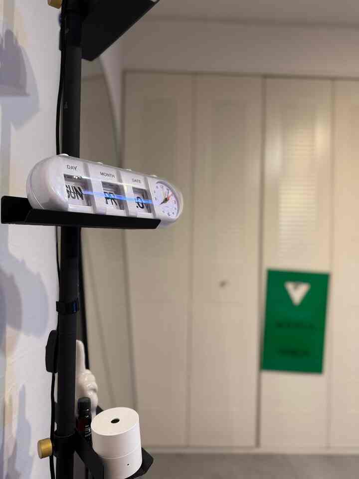 ブラックのテンションロッドに設置された白いアナログ兼カレンダー時計と、背景にぼかされたベージュ色のシャッターがあるシンプルなインテリア