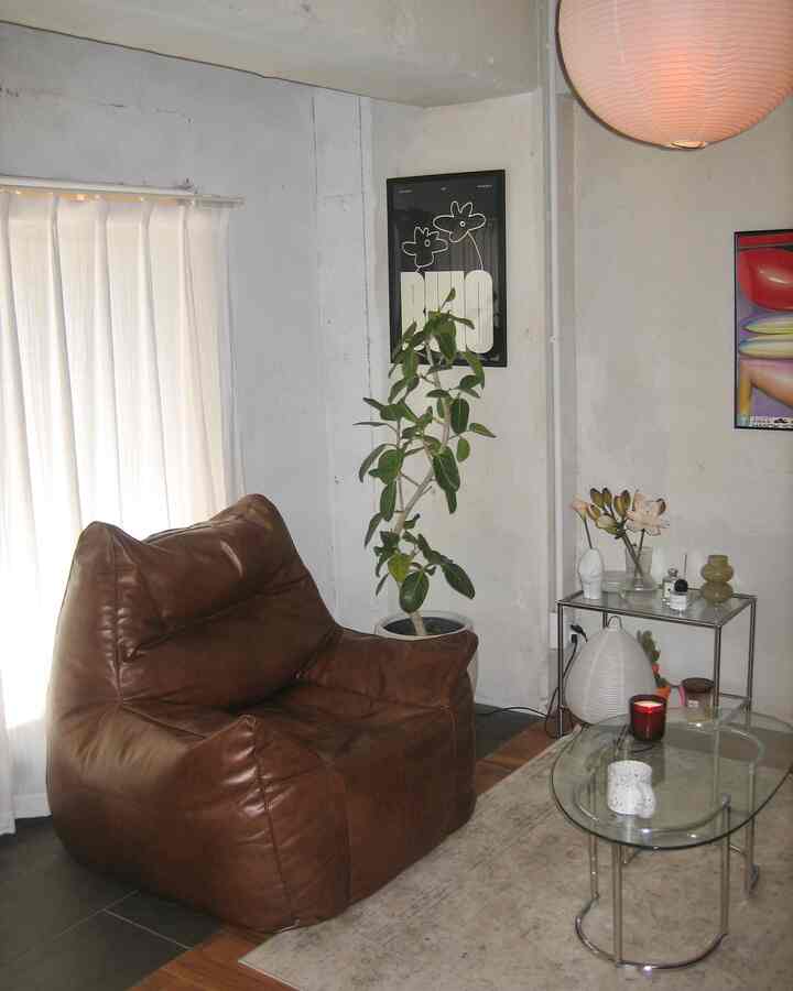 Natural living room with white walls and curtains, brown leather bean bag sofa, and glass low table featured in a cozy single household space