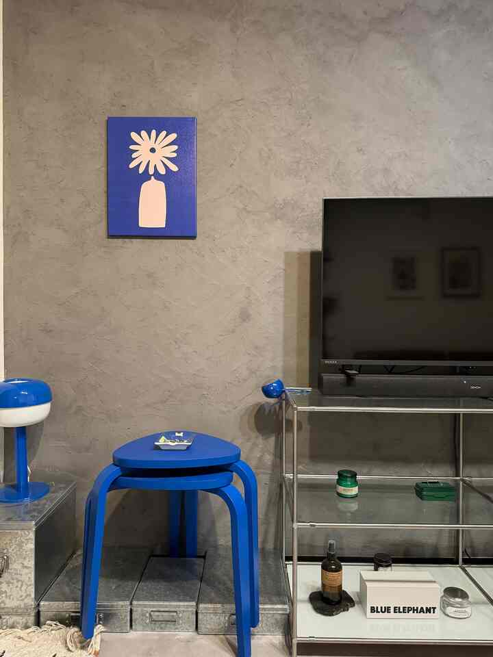 グレーのテクスチャ壁とメタルラック、鮮やかな青のスツールとテーブルランプが配置されたモダンなリビングコーナー