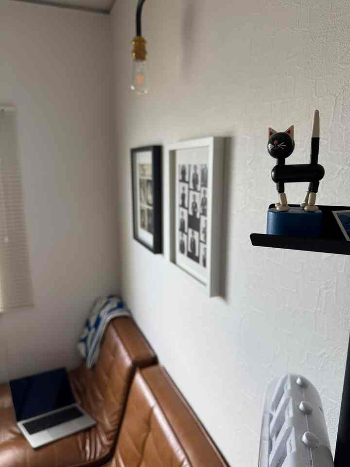 White-walled home office space featuring a brown leather sofa with blue patterned blanket, wall art, and a small wooden cat figurine creating a cozy atmosphere