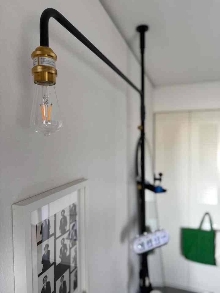 白い壁と黒いテンションロッド、ゴールドのジョイントが特徴的な壁照明が設置されたワンルームの額縁のある空間