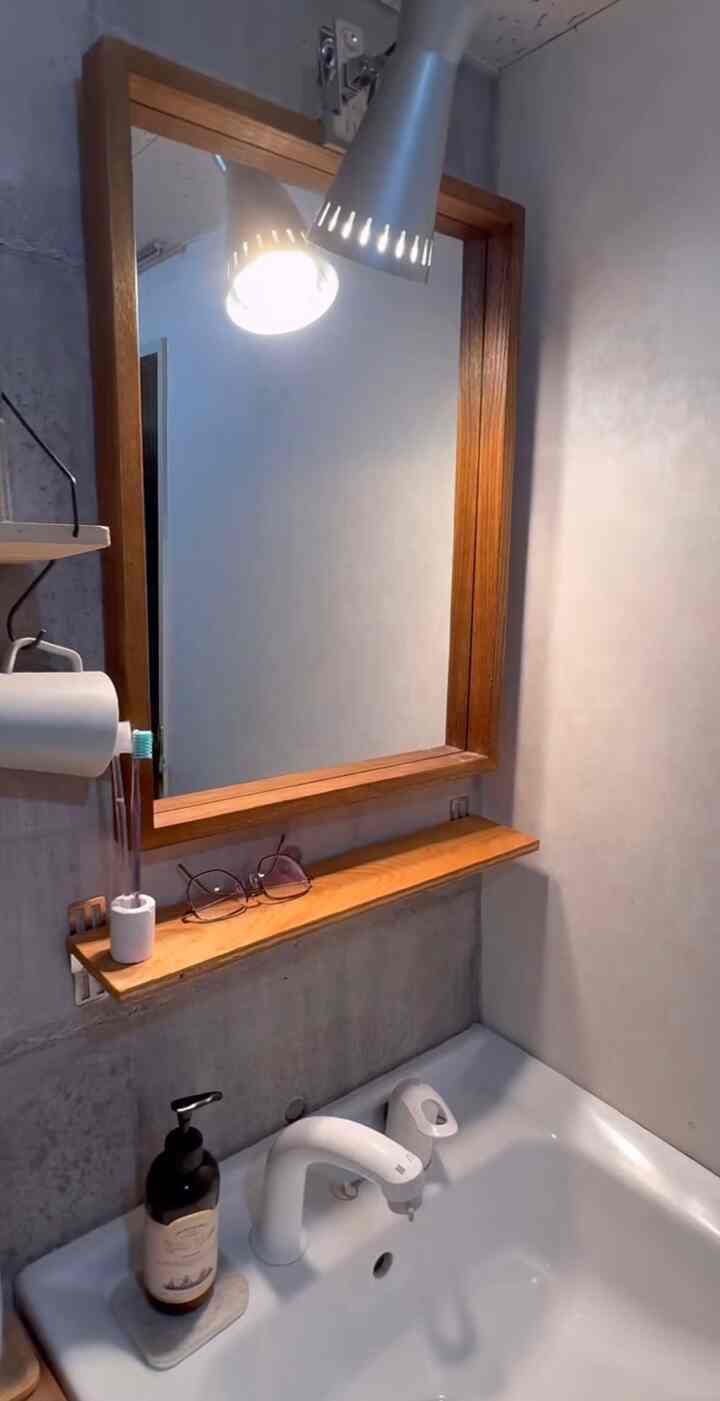 ナチュラルな木目調が際立つバスルーム空間、木製フレームの鏡と小さな棚、シルバーの照明が配置された落ち着いたインテリア