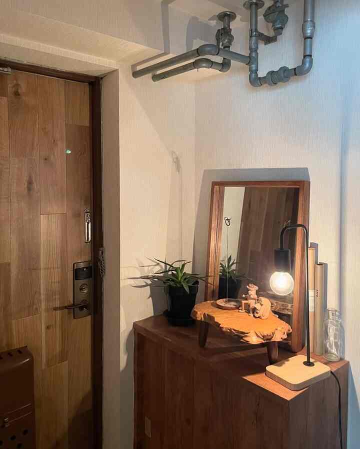 温かみのある木目調と白い壁の玄関、木製の鏡とテーブルランプ、植物が配置され、落ち着いた雰囲気のインテリア。