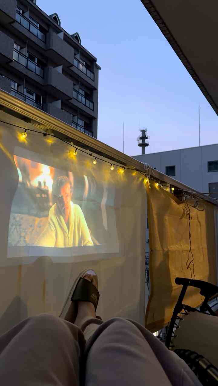 白いカーテンと黄色いストリングライトがあるベランダの空間、リクライニングチェアとプロジェクターで演出された雰囲気のある屋外シネマ