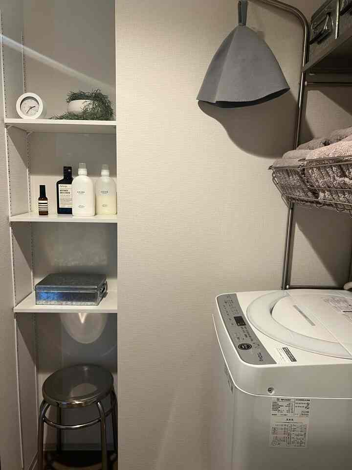 グレーと白の落ち着いた色調のバスルーム、メタルスツールと収納棚、洗濯機とサウナ帽が配置された清潔感のあるインテリア