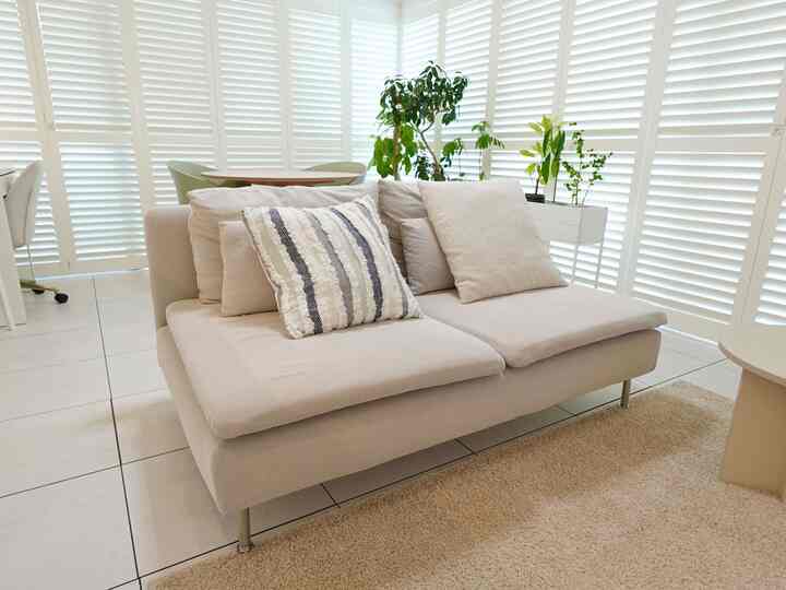 明るいベージュ調のリビング、IKEAソファと白いウッドシャッター、植物が配置され、柔らかく広々とした雰囲気のインテリア