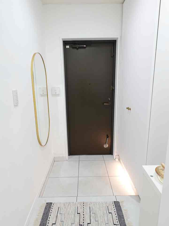 白い壁と靴箱が配置されたコンパクトな玄関、ゴールドフレームの楕円形ミラーとセンサーライトが印象的なモダンな空間