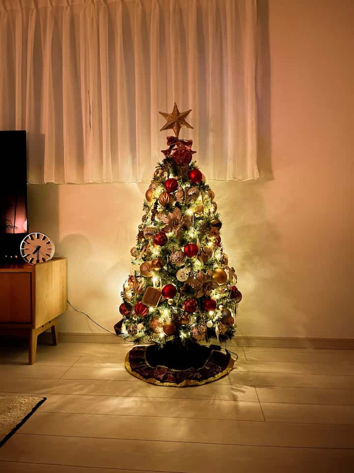 暖かい照明に照らされた白いカーテンと木の床があるリビングの中央に、赤とゴールドのオーナメントが飾られたクリスマスツリーが立っている。