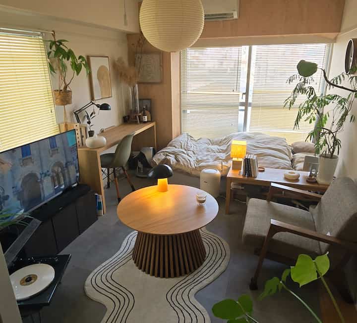 ナチュラルな色調のリビング兼寝室、丸テーブルとペンダントライト、植物や木目調の家具が配置された雰囲気のあるインテリア。