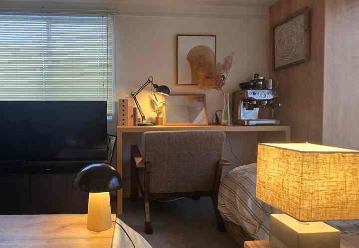 ベージュと木目調のホームオフィス兼寝室、デスクとアームチェア、テーブルランプとコーヒーマシンが配置されたナチュラルなインテリア