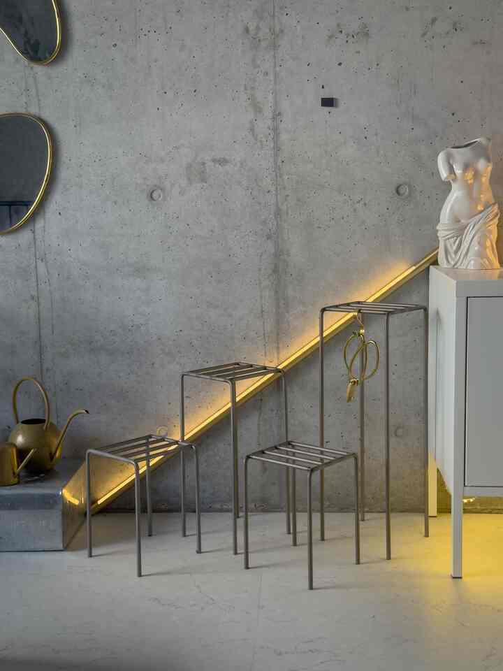 コンクリートの壁と白い床、ゴールドの照明がアクセントのモダンな空間に金属製のプラントスタンドセットが配置されている。