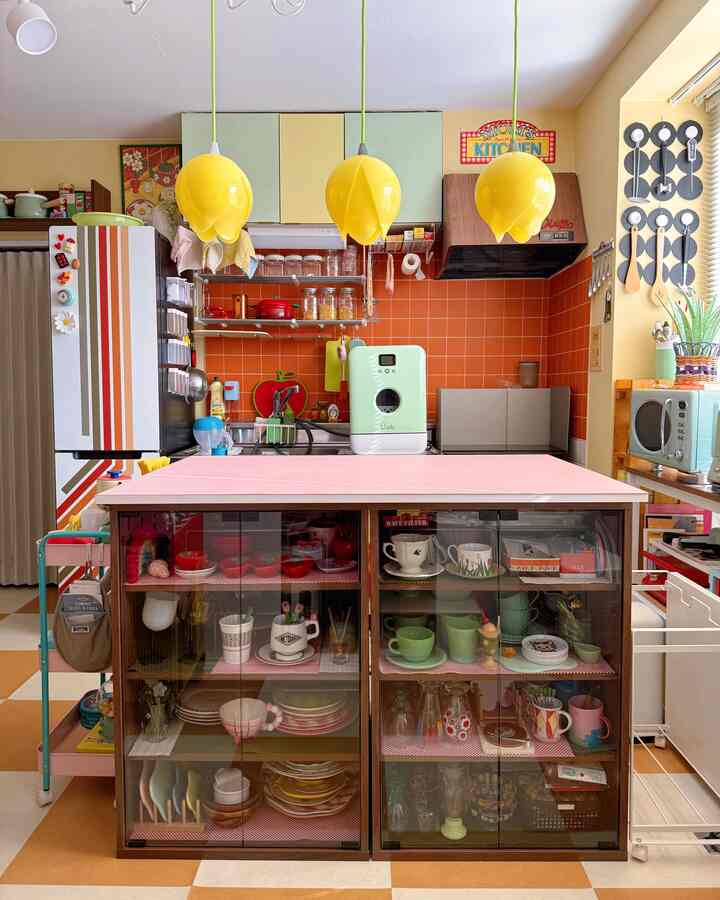 イエローの照明とモジュラー収納キャビネットがあるレトロなキッチン、ピンクの天板とオレンジのタイル壁が印象的な明るい空間