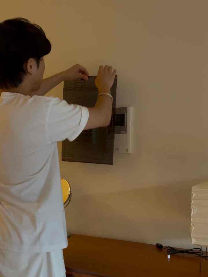 ナチュラルな色調の壁と木製のデスクを備えた空間、中央の壁に取り付けられたインターホンをダークカラーのすだれマットで覆い、実用的で整った雰囲気のインテリア