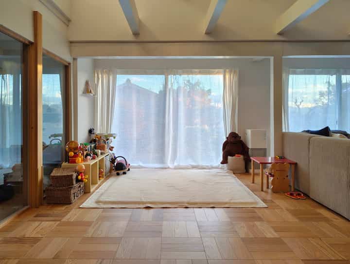 明るい白いカーテンとベージュのソファが配置されたナチュラルな色調のリビング、子供のおもちゃと小さなテーブルがある遊びスペースを含む雰囲気のあるインテリア
