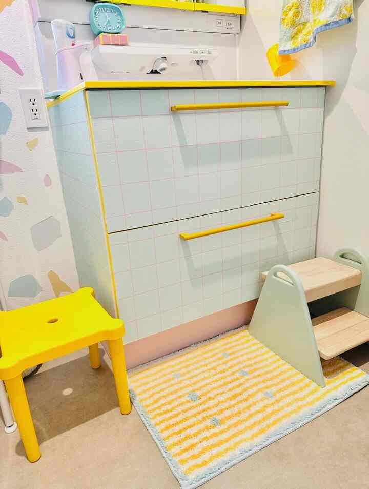 明るい黄色とパステル調の子供部屋、スツールとキッズステップ、ストライプ柄のラグが配置された柔らかい雰囲気のインテリア