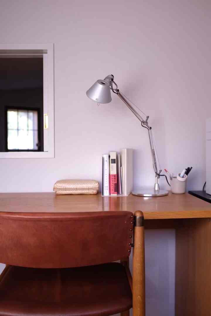 白い壁とナチュラルウッドのデスク、ブラウンレザーのオフィスチェア、シルバーのテーブルランプが配置された2階のワークスペース