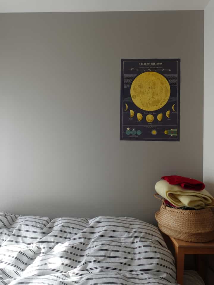 白を基調としたコンパクトな6畳の寝室、ストライプ柄のベッドと木製のナイトテーブル、壁に掲げられた月のチャートポスターが配置された落ち着いたインテリア