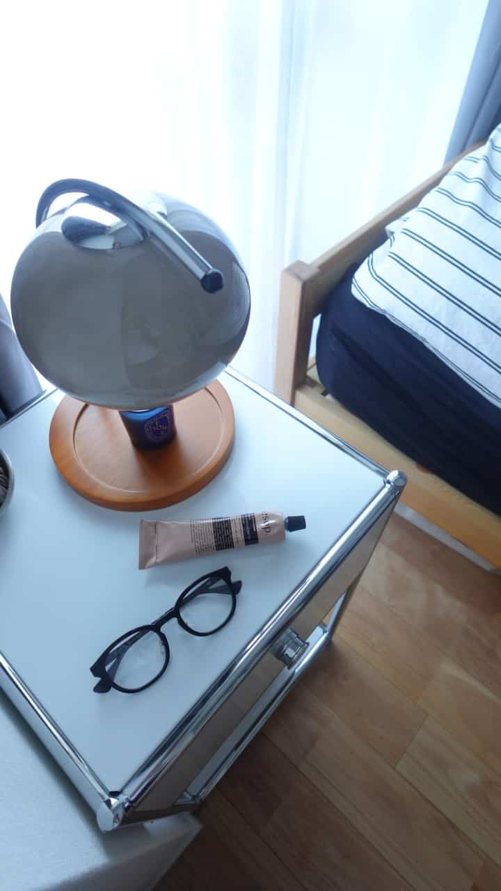 ホワイトと木目調の寝室、ナイトテーブルの上に黒い眼鏡とチューブ、丸いランプが置かれた清潔感のあるインテリア