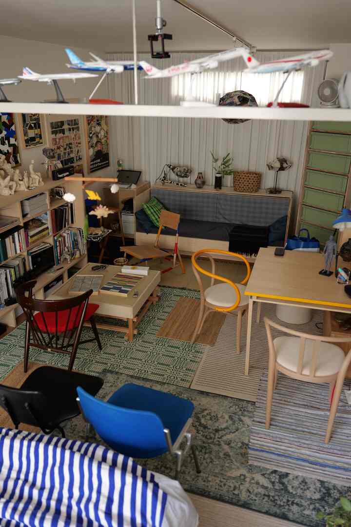 ナチュラルな色調とミッドセンチュリーモダンスタイルのワンルームリビング、多様な椅子と本棚、ラグが配置された温かみのある空間