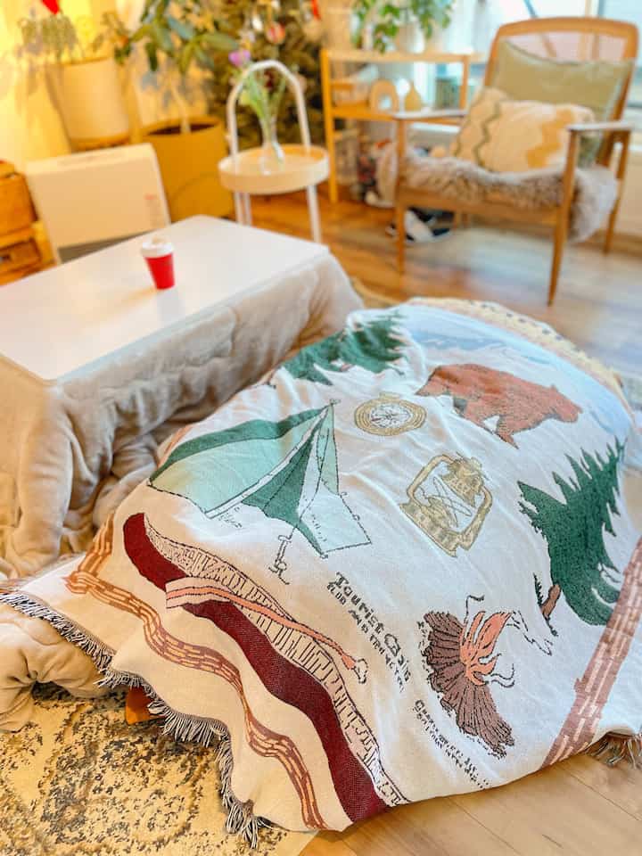 温かみのあるウッドトーンとナチュラルな装飾が調和した冬のリビング、中央にキャンプ柄のスローをかけたこたつが置かれた印象的な空間