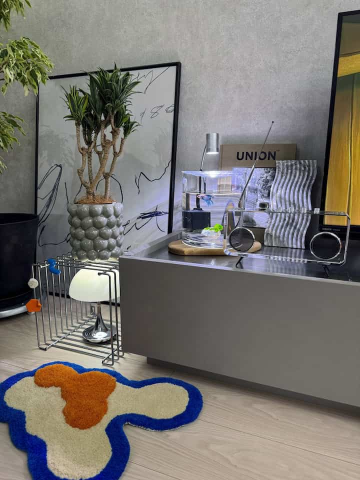 グレーの壁を背景にしたナチュラルトーンのリビング、中央左に大きな植物、中央右に水槽と多様な装飾品が配置された空間