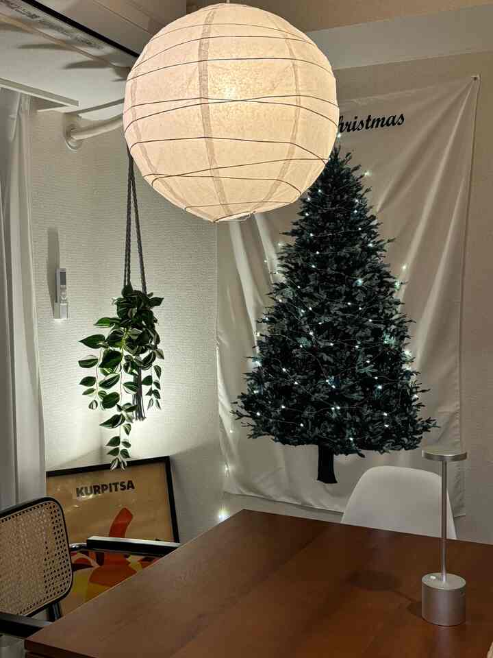 ナチュラルな色調のダイニングルーム、中央に木製のダイニングテーブルとラタンチェアが配置され、壁にはクリスマスツリーのタペストリーとストリングライトが飾られた暖かみのある空間