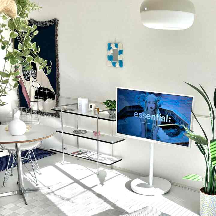 White-toned living room featuring a round dining table with wire chairs, TV stand, and shelving unit in a minimal style