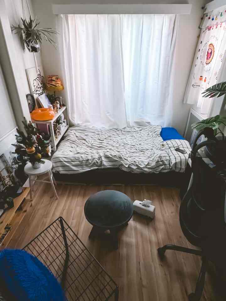 白い壁とカーテン、ブラウンの木目調の床が調和した狭いワンルーム寝室で、ベッド、スツール、デスクが配置された雰囲気のあるインテリア