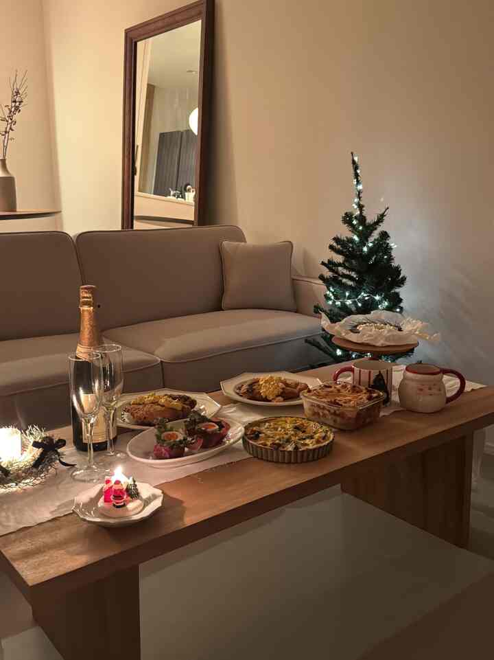 柔らかいベージュとブラウンの色調のリビング、アットホームなソファと小さなライトアップされたクリスマスツリーの横にある木製テーブルにホームパーティーの料理と装飾が配置された雰囲気のあるインテリア