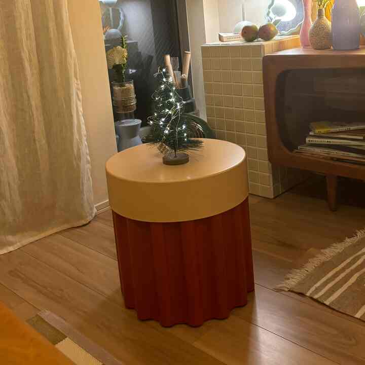 ベージュの壁とカーテンがあるリビング、中央に黄色い天板と赤みがかった台座の丸いコーヒーテーブルが置かれ、上には小さなLEDストリングライトの飾りがある。