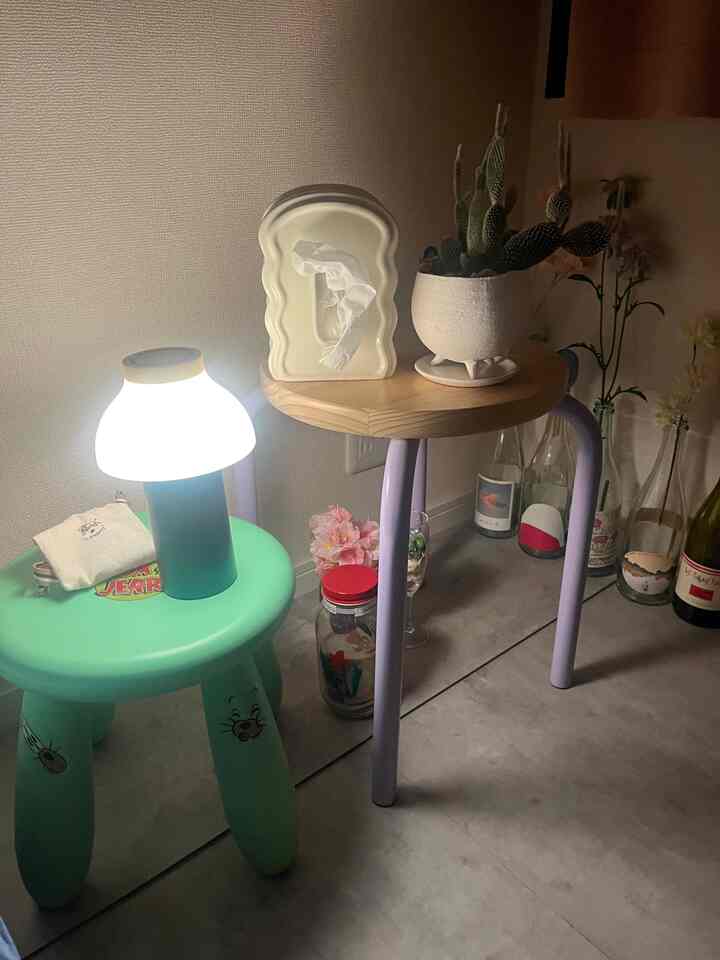 柔らかな照明の下、ミント色の子供用スツールと木製天板の紫色の脚のサイドテーブル、サボテンの植物が配置された居心地の良いミニマルな空間