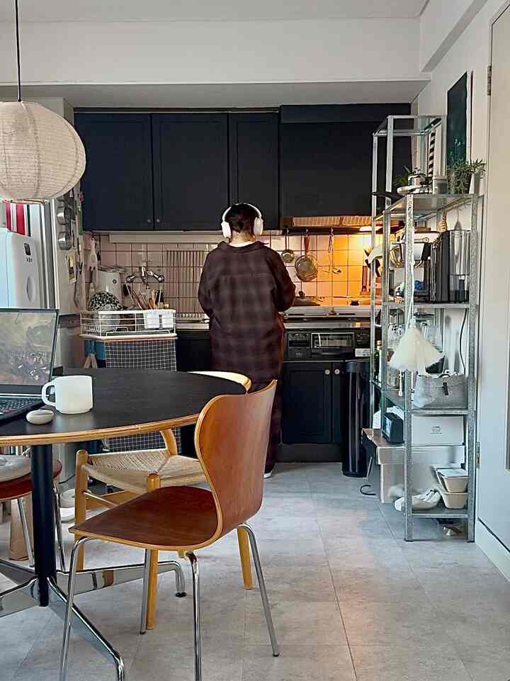 ブラックのキッチンとウッドトーンのダイニングチェアが配置されたナチュラルな1LDKキッチン・ダイニング室、料理中の人物と近くにくつろぐ猫が見える印象的な空間