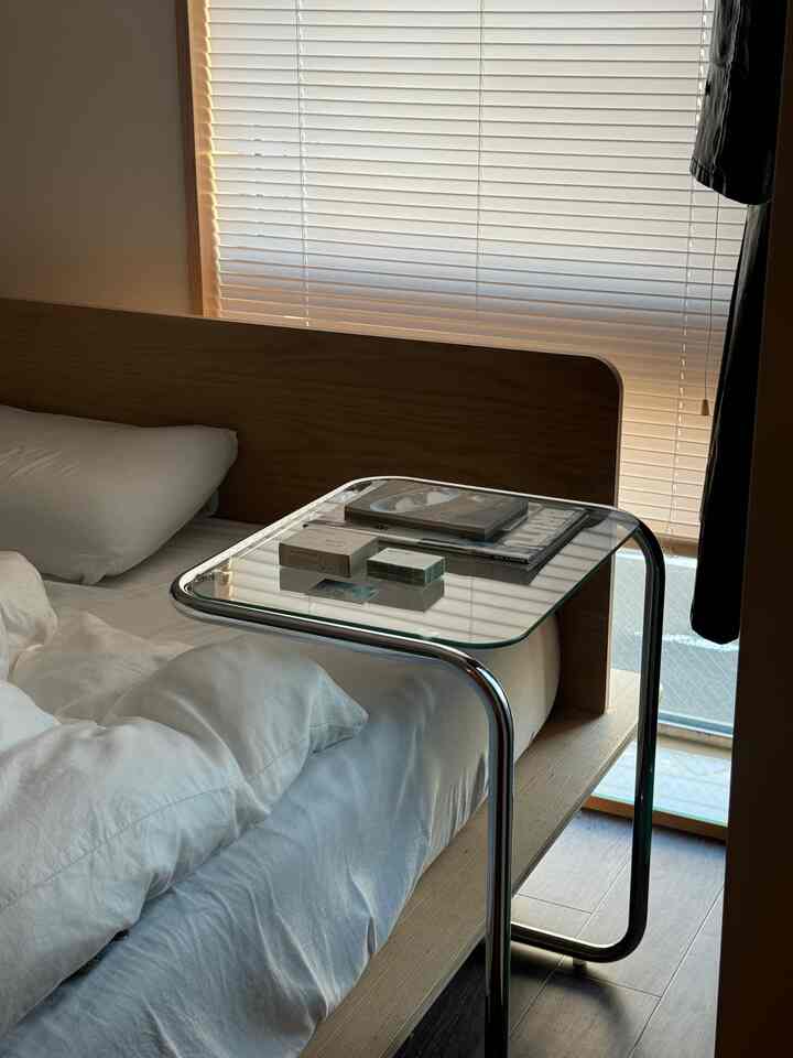 ナチュラルカラーとホワイトを基調とした寝室、ミッドセンチュリーモダンスタイルの透明なガラスのナイトテーブルが配置されたシンプルで洗練されたインテリア