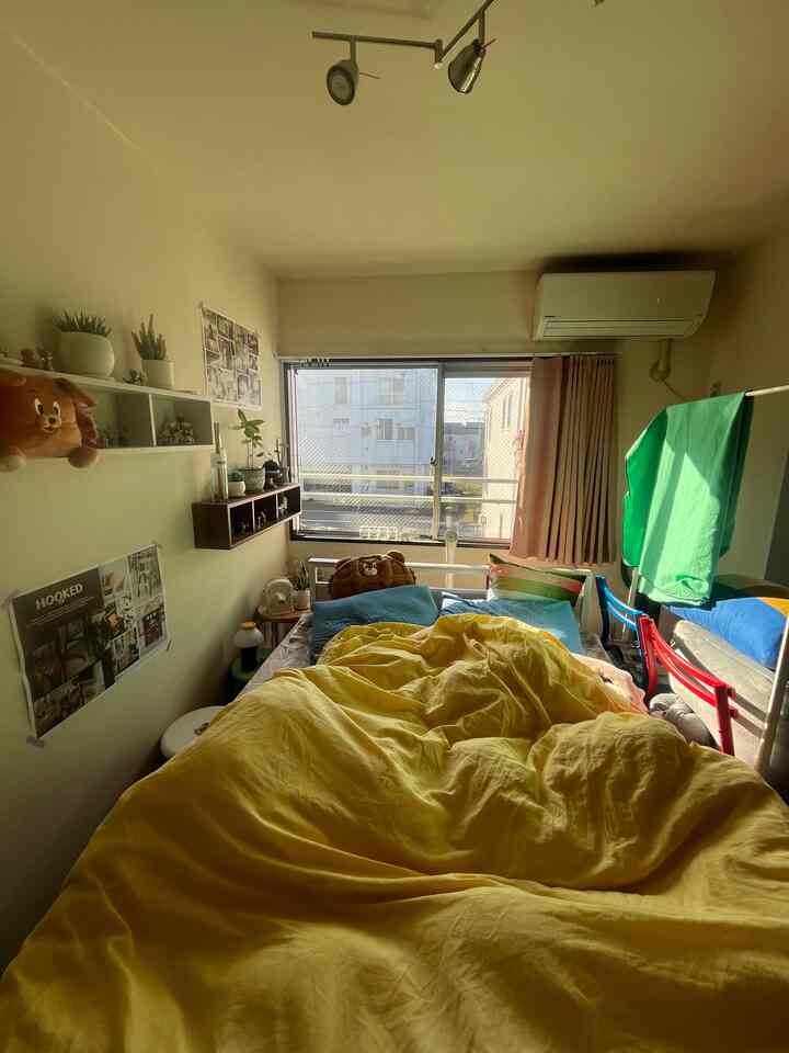 黄色い寝具が印象的な、壁掛け棚とカーテンが配置された自然光の入る居心地の良い小さな寝室