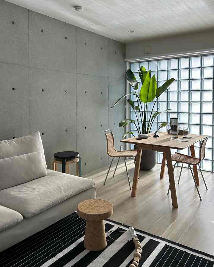 グレーのコンクリート壁と木目調の床が調和したモダンでナチュラルなリビング、コルクのスツールとダイニングチェアが配された清潔で明るい空間