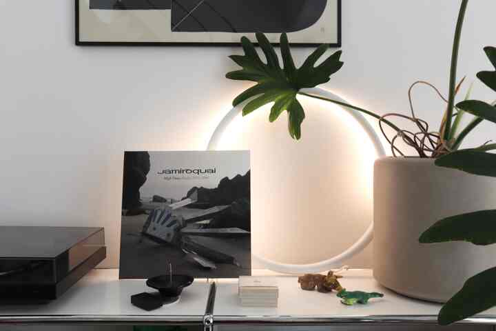 白いテーブルの上にレコードプレーヤーとジャミロクアイのアルバムカバー、壁に照明と鉢植えが配置された雰囲気のある空間